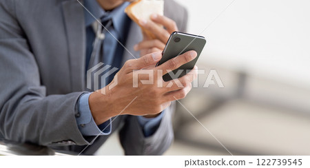 Close up of a young Asian businessman holding a smartphone and a snack in a modern office environment. Close up of a young Asian businessman holding a smartphone and a snack in a modern office environment. 122739545