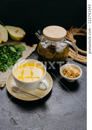 Pear and mint herbal tea in cup with glass teapot, fresh ingredients, and sugar cubes on dark table 122741043