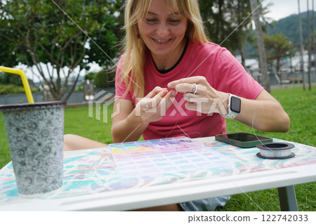 Young woman beading colorful bracelets outdoors in a park Young woman beading colorful bracelets outdoors in a park 122742033