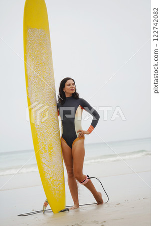 A woman in a stylish wet suit confidently holds a surfboard on the beach A woman in a stylish wet suit confidently holds a surfboard on the beach 122742082