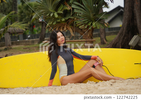 Woman in stylish wetsuit posing confidently with surfboard on beach 122742225