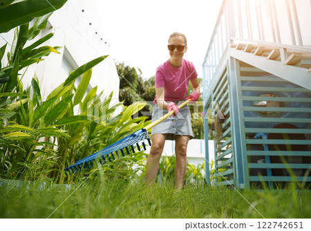 A woman raking the lawn at the backyard of her house  122742651