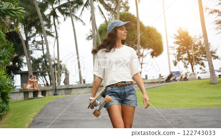 A woman is walking at the city park while carrying a skateboard in her hands 122743033