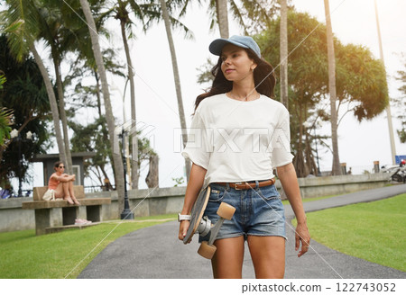A woman is walking at the city park while carrying a skateboard in her hands 122743052
