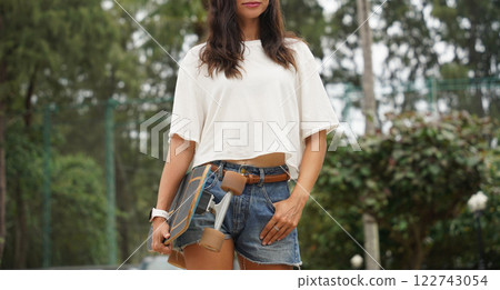 A woman is walking at the city park while carrying a skateboard in her hands A woman is walking at the city park while carrying a skateboard in her hands 122743054