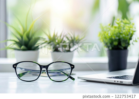 Close up of eyeglasses on work desk with laptop at business workplace. Black glasses on white table, copy space. Empty space for text. Keyboard, notebook, eye glasses and plants. Office accessories 122753417