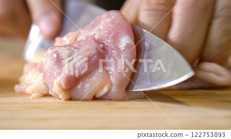 A chef using a kitchen knife slices uncooked chicken meat on a chopping board, the close up view of preparing food ingredients. A chef using a kitchen knife slices uncooked chicken meat on a chopping board, the close up view of preparing food ingredients. 122753893