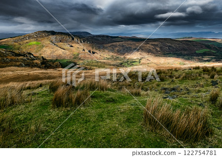 A landscape of varied terrain featuring hills, valleys, and grassy plains. The sky is mostly overcast with some breaks where sunlight penetrates. Vegetation is predominantly dry grass and shrubs. 122754781