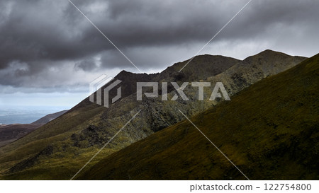 Jagged mountain ridges under a dramatic, cloudy sky, visible lower terrain with some greenery. Jagged mountain ridges under a dramatic, cloudy sky, visible lower terrain with some greenery. 122754800