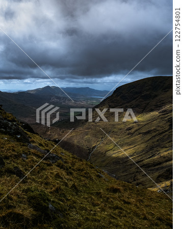 High angle shot of a rugged landscape.  Shadows dominate the foreground slopes, while sunlight touches distant peaks and a winding stream. 122754801