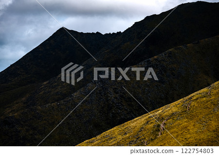 Multiple dark mountain summits, layered and steep, with a stormy sky above and glimpses of lower land. 122754803