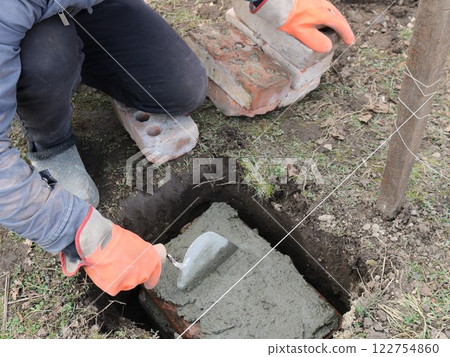 worker in orange gloves laying and leveling a layer of fresh cement with a trowel in a hole in the ground on a block with a string stretched nearby as a level, preparing for foundation work 122754860