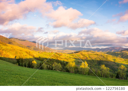 mountain landscape in spring. trees behind green meadow at sunset. countryside scenery with rolling hills. sky with clouds in evening light picturesque destination for outdoor adventure 122756180