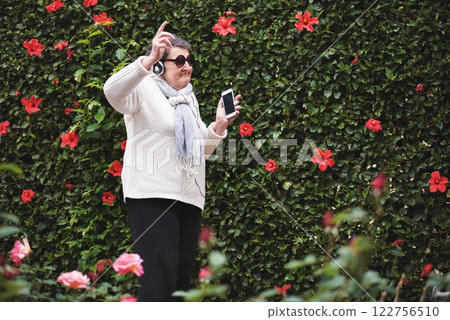 Senior woman, headphones and dancing in outdoors, smartphone and streaming radio or playlist. Elderly person, flower wall and rock or pop music in retirement, garden and freedom or connection to tech Senior woman, headphones and dancing in outdoors, smartphone and streaming radio or playlist. Elderly person, flower wall and rock or pop music in retirement, garden and freedom or connection to tech 122756510