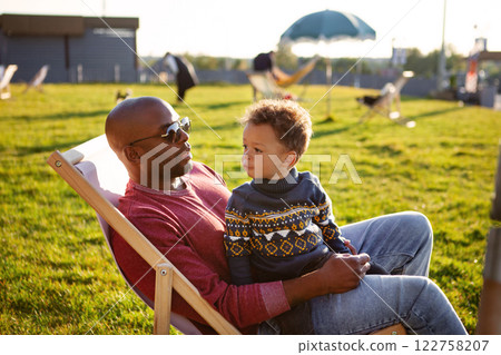 Father and young son spending quality time together outdoors on sunny day, sitting on lounge chair in park during golden hour surrounded by green grass, wearing cozy stylish sweaters. Dad and child 122758207