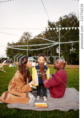 Family picnic in park. Mom, dad and child enjoying delicious snacks and quality time together surrounded by nature and festive decorations under evening sky. Mother, Father and son sitting on grass 122758226
