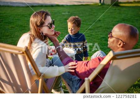 Happy Family holiday, Mother and Father With Child Enjoying Quality Time Together, Relaxing On Lounge Chairs On green Grass In Park On Warm And Sunny Day. 122758281
