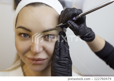 The hand of a make-up artist in black gloves applies a yellow eyebrow paste to the eyebrow. Professional stylish permanent makeup, eyebrow tinting. Close up. The hand of a make-up artist in black gloves applies a yellow eyebrow paste to the eyebrow. Professional stylish permanent makeup, eyebrow tinting. Close up. 122760479