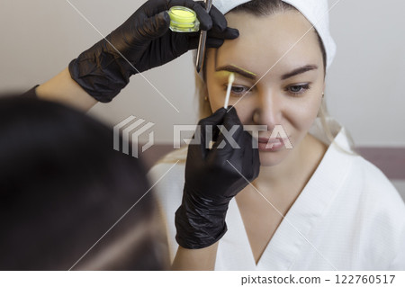 The hand of a make-up artist in black gloves applies a yellow eyebrow paste to the eyebrow. Professional stylish permanent makeup, eyebrow tinting. Close up. The hand of a make-up artist in black gloves applies a yellow eyebrow paste to the eyebrow. Professional stylish permanent makeup, eyebrow tinting. Close up. 122760517