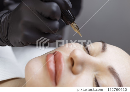 Close-up of a woman, in a beauty salon with her eyes closed, a cosmetologist makes permanent eyeliner makeup. Professional in black gloves and with a special too Close-up of a woman, in a beauty salon with her eyes closed, a cosmetologist makes permanent eyeliner makeup. Professional in black gloves and with a special too 122760610