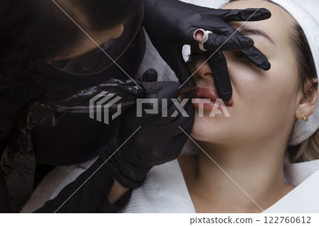 Permanent makeup for nude lips. Cosmetologist in the salon performs a tattoo procedure with a tattoo machine close-up 122760612
