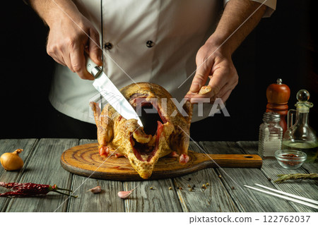 A skilled chef carves a raw duck carcass that rests on a rustic wooden board. Fresh ingredients and spices surround the scene, hinting at the passion for culinary art in a cozy kitchen 122762037