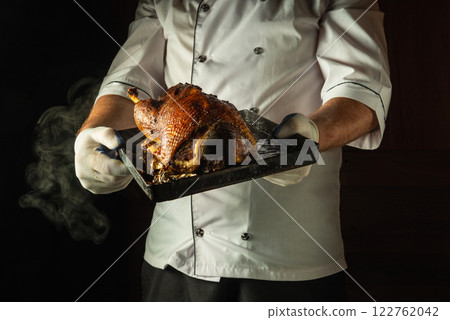 In a dimly lit kitchen, a skilled chef proudly holds a roasted duck on a tray. The rich golden-brown skin glistens, inviting the senses to indulge in this exquisite culinary creation In a dimly lit kitchen, a skilled chef proudly holds a roasted duck on a tray. The rich golden-brown skin glistens, inviting the senses to indulge in this exquisite culinary creation 122762042