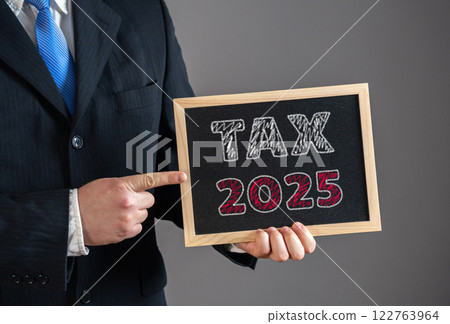 A businessman points to a sign that says Tax 2025. Upcoming tax policies, financial planning, fiscal regulations, corporate taxation. Tax deadlines, government levies, financial obligations. A businessman points to a sign that says Tax 2025. Upcoming tax policies, financial planning, fiscal regulations, corporate taxation. Tax deadlines, government levies, financial obligations. 122763964
