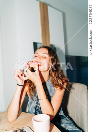 A cheerful young woman bites into a pink-frosted donut topped with sprinkles, enjoying a sweet treat A cheerful young woman bites into a pink-frosted donut topped with sprinkles, enjoying a sweet treat 122764085