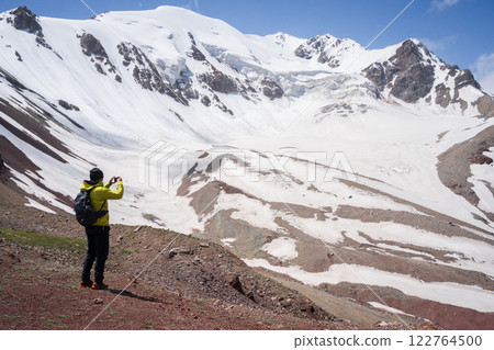 Male hiker taking photo of alpine landscape with snowy peaks, glacier in Pamir Mountains, Kyrgyzstan Male hiker taking photo of alpine landscape with snowy peaks, glacier in Pamir Mountains, Kyrgyzstan 122764500