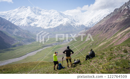 Hikers enjoying views on green alpine valley with massive glaciers of Pamir Mountains, Kyrgyzstan Hikers enjoying views on green alpine valley with massive glaciers of Pamir Mountains, Kyrgyzstan 122764509