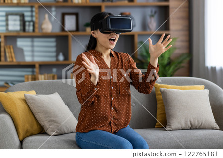 Excited asian woman wearing virtual reality headset experiencing immersive digital content in cozy living room. Expression shows amazement and engagement with modern VR technology and lifestyle. Excited asian woman wearing virtual reality headset experiencing immersive digital content in cozy living room. Expression shows amazement and engagement with modern VR technology and lifestyle. 122765181