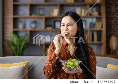 Asian woman eating salad with dissatisfied expression, contemplating diet. Seated on couch in contemporary interior setting, representing healthy lifestyle choices. Concept of wellness journey. Asian woman eating salad with dissatisfied expression, contemplating diet. Seated on couch in contemporary interior setting, representing healthy lifestyle choices. Concept of wellness journey. 122765191