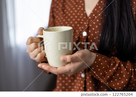 Close-up of asian woman holding mug, displaying comfort, warmth, and relaxation. Hands cradling neutral-colored cup, wearing red polka dot blouse, suggesting cozy, calm atmosphere. Close-up of asian woman holding mug, displaying comfort, warmth, and relaxation. Hands cradling neutral-colored cup, wearing red polka dot blouse, suggesting cozy, calm atmosphere. 122765204