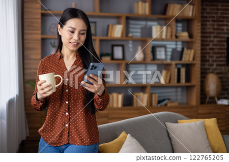 Asian woman holds smartphone and coffee, standing by cozy couch with bookshelf. Relaxed indoor setting, reading message on phone, enjoying leisure time at home. Asian woman holds smartphone and coffee, standing by cozy couch with bookshelf. Relaxed indoor setting, reading message on phone, enjoying leisure time at home. 122765205