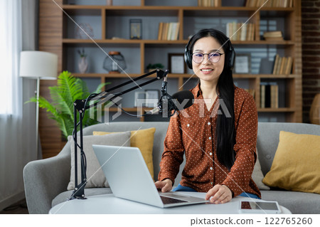 Asian woman hosting podcast in home studio, seated on sofa with laptop and microphone. Headphones on, conveying technology and communication in cozy living space with home office vibes. 122765260