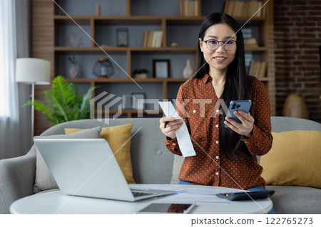 Asian woman managing finances at home with smartphone and laptop. She holds receipt, showing engagement in budgeting. Comfortable domestic setting enhances productive atmosphere. Asian woman managing finances at home with smartphone and laptop. She holds receipt, showing engagement in budgeting. Comfortable domestic setting enhances productive atmosphere. 122765273