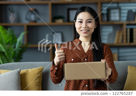 Asian woman with long hair and dotted shirt holding cardboard box, giving thumbs up on sofa. Captures happiness, success, home comfort, and positivity Asian woman with long hair and dotted shirt holding cardboard box, giving thumbs up on sofa. Captures happiness, success, home comfort, and positivity 122765287