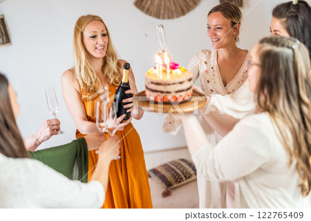 Birthday surprise party. Group of cheerful female friends celebrating personal success stories. Smiling girls toasting with glasses of champagne one holding a birthday cake with candles to blow. Birthday surprise party. Group of cheerful female friends celebrating personal success stories. Smiling girls toasting with glasses of champagne one holding a birthday cake with candles to blow. 122765409