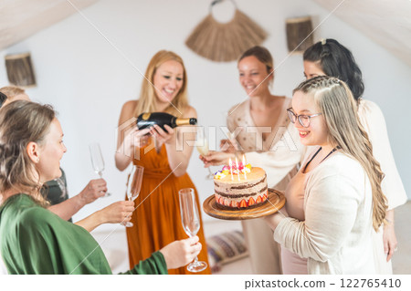 Birthday surprise party. Group of cheerful female friends celebrating personal success stories. Smiling girls toasting with glasses of champagne one holding a birthday cake with candles to blow. 122765410