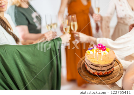 Birthday surprise party. Group of cheerful female friends celebrating personal success stories. Smiling girls toasting with glasses of champagne one holding a birthday cake with candles to blow. 122765411