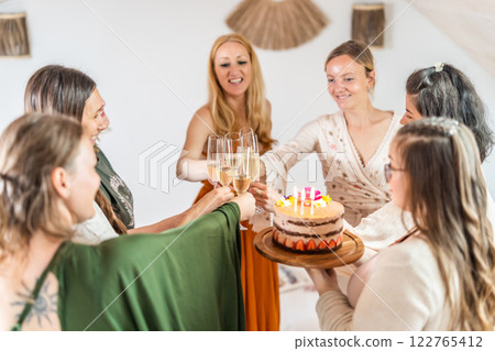 Birthday surprise party. Group of cheerful female friends celebrating personal success stories. Smiling girls toasting with glasses of champagne one holding a birthday cake with candles to blow. 122765412