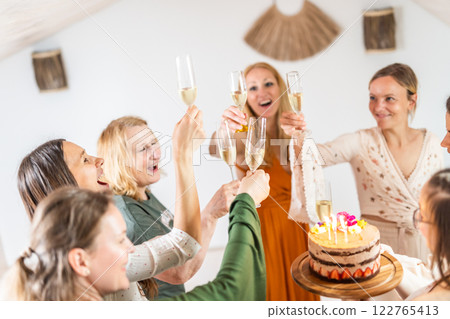 Birthday surprise party. Group of cheerful female friends celebrating personal success stories. Smiling girls toasting with glasses of champagne one holding a birthday cake with candles to blow. 122765413