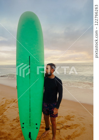 Young bearded man with surfboard standing near a beach. Man with surfing board outdoors on a summer day. Young bearded man with surfboard standing near a beach. Man with surfing board outdoors on a summer day. 122766263