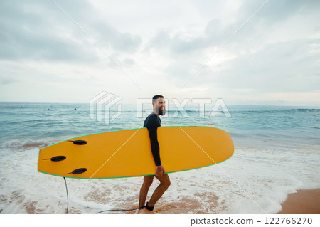 Young handsome male surfer with a surfboard is enjoying a view while walking a sandy beach at sea. Summer, vacation, ocean Young handsome male surfer with a surfboard is enjoying a view while walking a sandy beach at sea. Summer, vacation, ocean 122766270