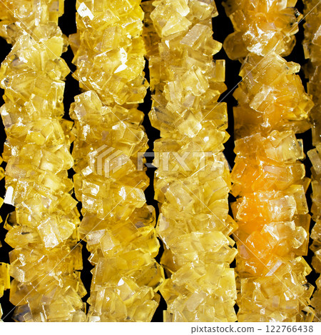 The sticks are made of sugar and are arranged in a line. A row of yellow crystals are lined up on a black surface. The crystals are all different sizes and shapes 122766438