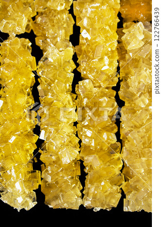 The sticks are made of sugar and are arranged in a line. A row of yellow crystals are lined up on a black surface. The crystals are all different sizes and shapes 122766439