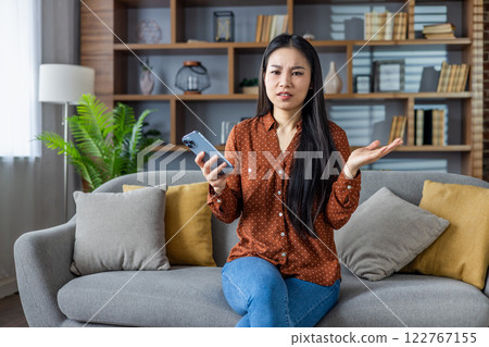 Asian woman seated on sofa holding smartphone looks confused and frustrated, expressing bewilderment and doubt. Comfortable home environment with bookshelves in background adds a cozy touch. Asian woman seated on sofa holding smartphone looks confused and frustrated, expressing bewilderment and doubt. Comfortable home environment with bookshelves in background adds a cozy touch. 122767155
