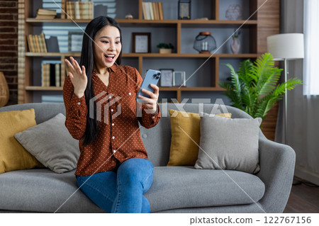 Asian woman celebrates online success, excited expression, holding smartphone. Sitting on cozy couch, modern living room environment. Positive vibes, home comfort, digital connection theme. Asian woman celebrates online success, excited expression, holding smartphone. Sitting on cozy couch, modern living room environment. Positive vibes, home comfort, digital connection theme. 122767156