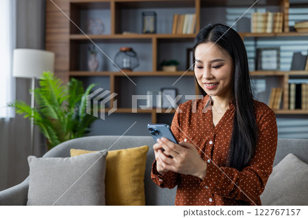 Asian woman smiling and using smartphone at home. Seated on sofa in modern living room, engaged in social media browsing or chatting. Captures moments of relaxation and connectivity. Asian woman smiling and using smartphone at home. Seated on sofa in modern living room, engaged in social media browsing or chatting. Captures moments of relaxation and connectivity. 122767157
